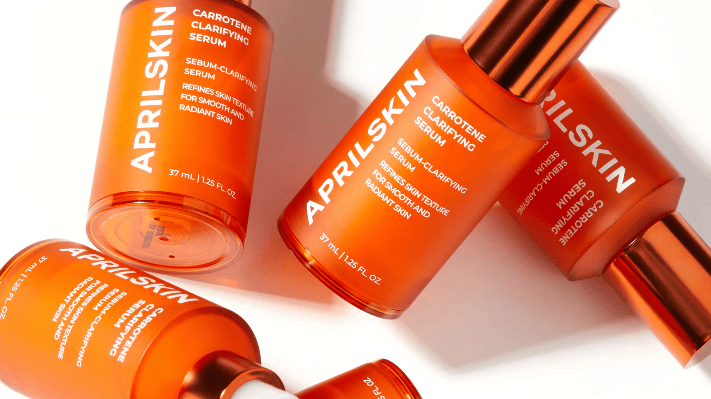 Bilden visar ett sortiment av APRILSKIN hudvårdsprodukter, specifikt deras "Carrotene Clarifying Serum." Flaskorna är enhetligt utformade med en iögonfallande orange färg, vilket speglar ingrediensen morot som är känd för sina hudklarande egenskaper.