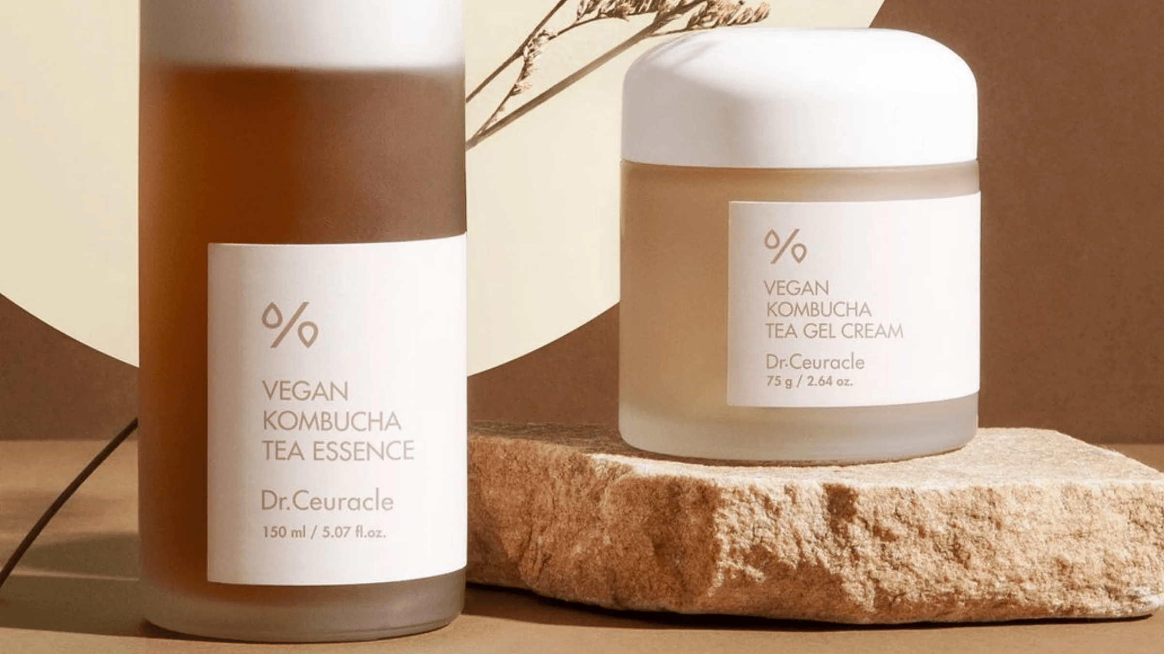 Dr. Ceuracle Vegan Kombucha Tea Essence och Tea Gel Cream i eleganta frostad glasförpackning, placerade på en stenplatta med naturlig belysning och jordnära bakgrund.
