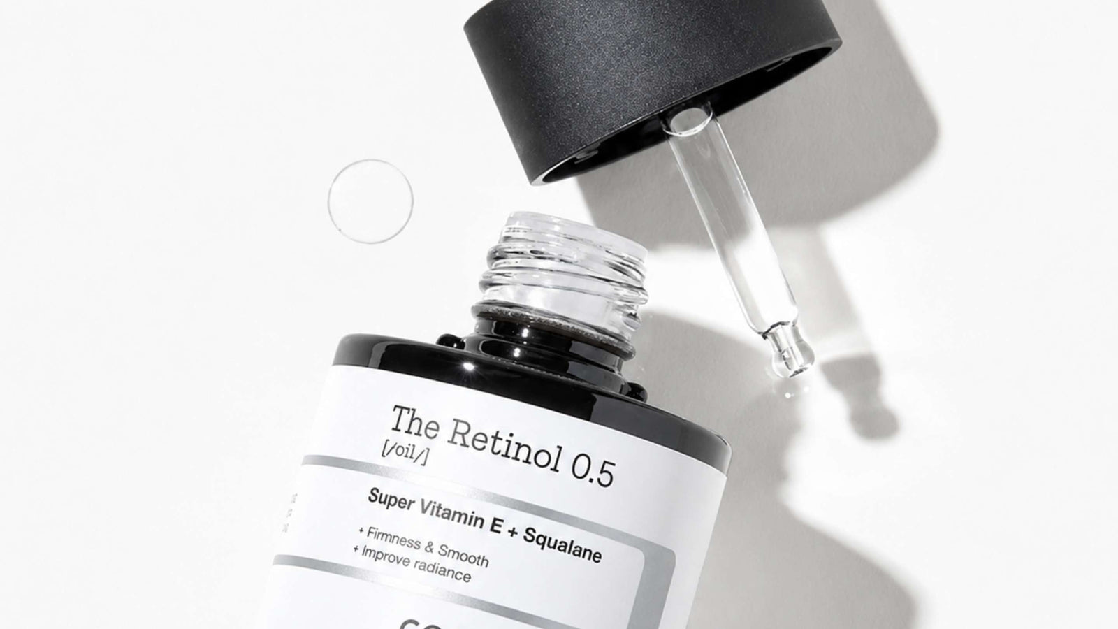 En droppflaska med COSRX The Retinol 0.5 olja ligger på sidan med pipetten droppande ut produkt, i solljus mot en grön bakgrund.