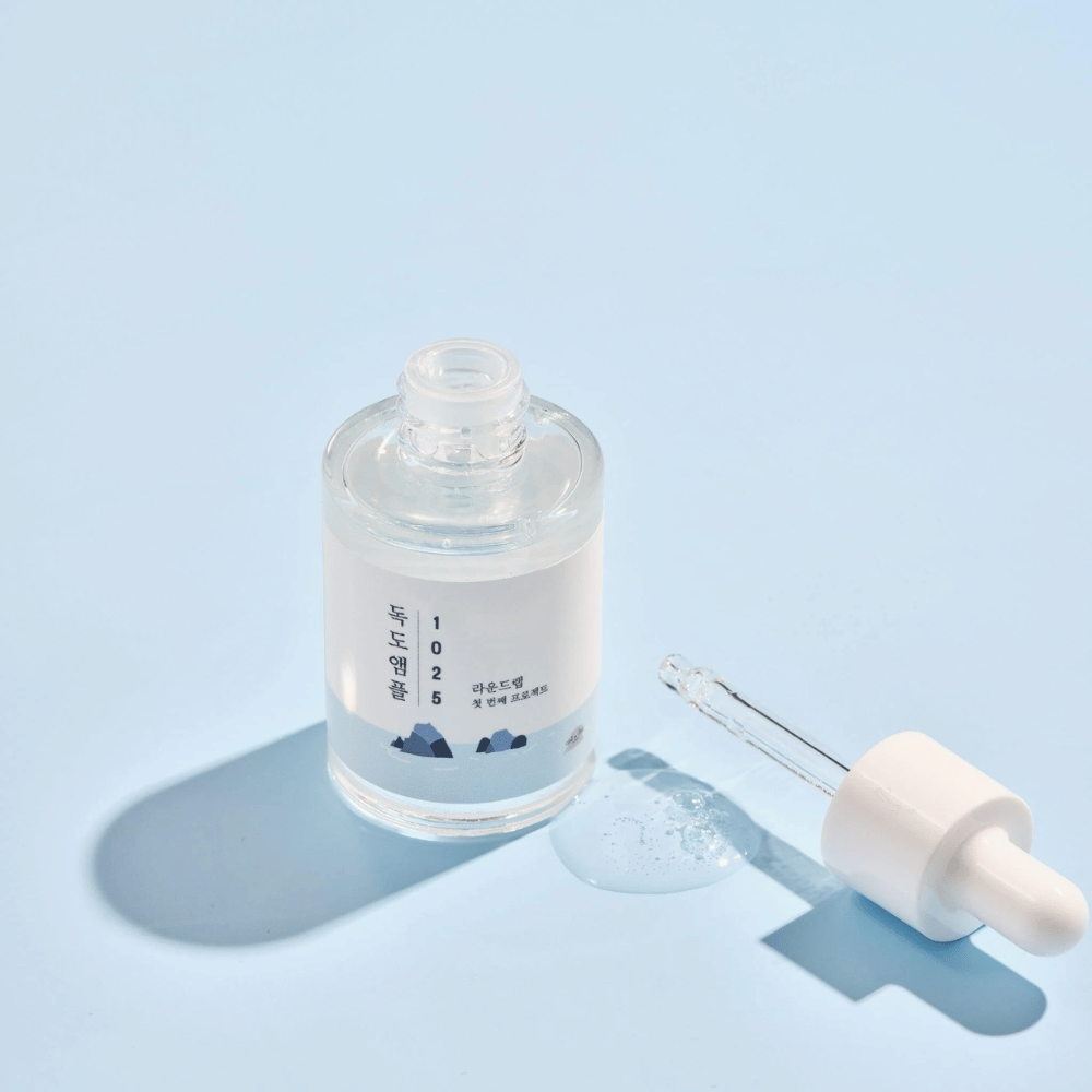 Eine offene Flasche von 1025 Dokdo Ampoule mit einer Pipette daneben, vor einem blauen Hintergrund, der einen weichen Schatten erzeugt.
