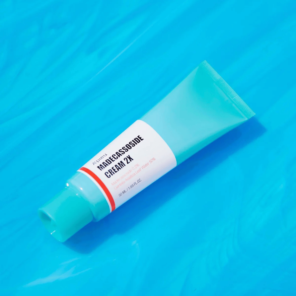 Eine Tube A'PIEU Madecassoside Cream 2X liegt diagonal über einem blauen Hintergrund mit wasserähnlichen Falten. Die Tube hat einen weißen Körper mit einem helltürkisen Deckel und ein rot bis orange gefärbtes Etikett, auf dem der Produktname und der aktive Inhaltsstoff Madecassoside 2% deutlich aufgedruckt sind. Auf dem Etikett steht auch die Menge, die 50 ml / 1,8 fl oz beträgt. Die helle und farbenfrohe Präsentation vermittelt ein frisches und energetisches Gefühl.