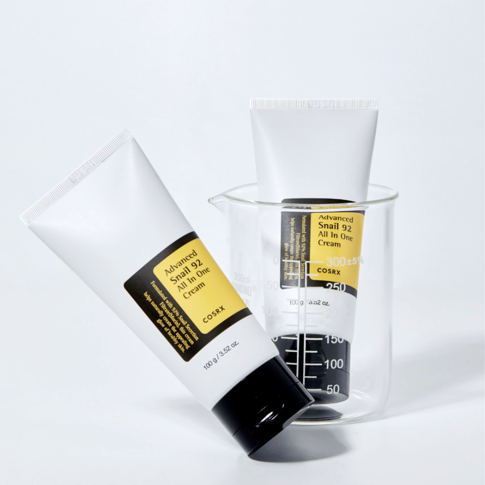 Eine Tube von COSRX Advanced Snail 92 All in One Cream liegend neben einem umgedrehten, durchsichtigen Messzylinder auf einem weißen Hintergrund.