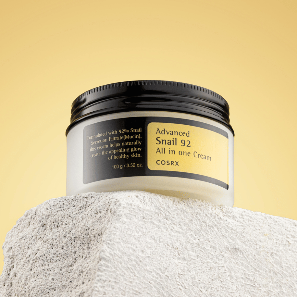 Eine Dose von COSRX Advanced Snail 92 All in one Cream auf einer steinigen Oberfläche mit gelbem Hintergrund platziert.