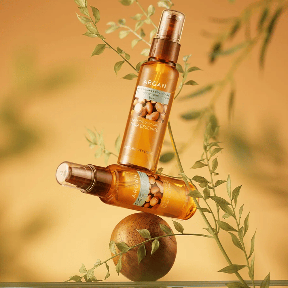 Zwei Flaschen Nature Republic Argan Essential Moisture Hair Oil zwischen grünen Blättern platziert – seidig glänzender Glanz für trockenes Haar.