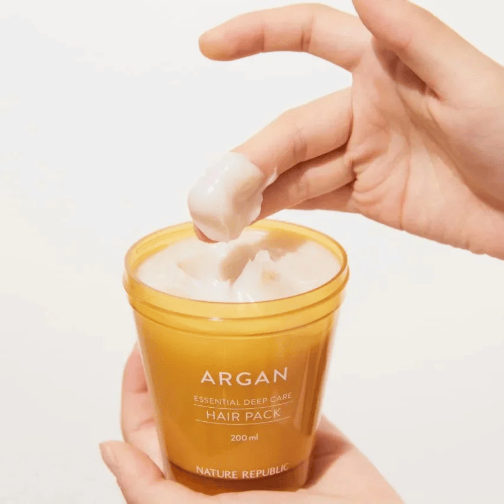 Hand nimmt die cremige Formel aus Nature Republic Argan Essential Moisture Repairing Hair Mask – intensive Pflege für trockenes Haar.