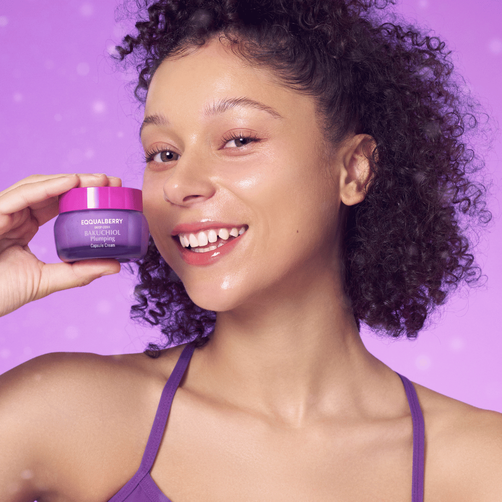 Frau mit strahlender Haut hält EQQUALBERRY Bakuchiol Plumping Capsule Cream und lächelt vor einem lila Hintergrund.