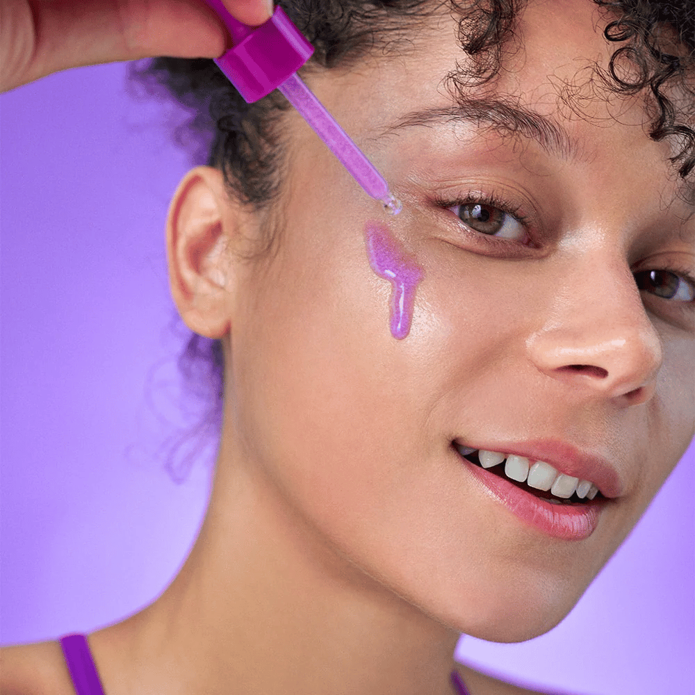 Lächelnde Frau trägt ein lila schimmerndes Serum mit einer Pipette auf die Haut auf. Strahlender Teint und lila Hintergrund, der zur Farbe des Produkts passt.