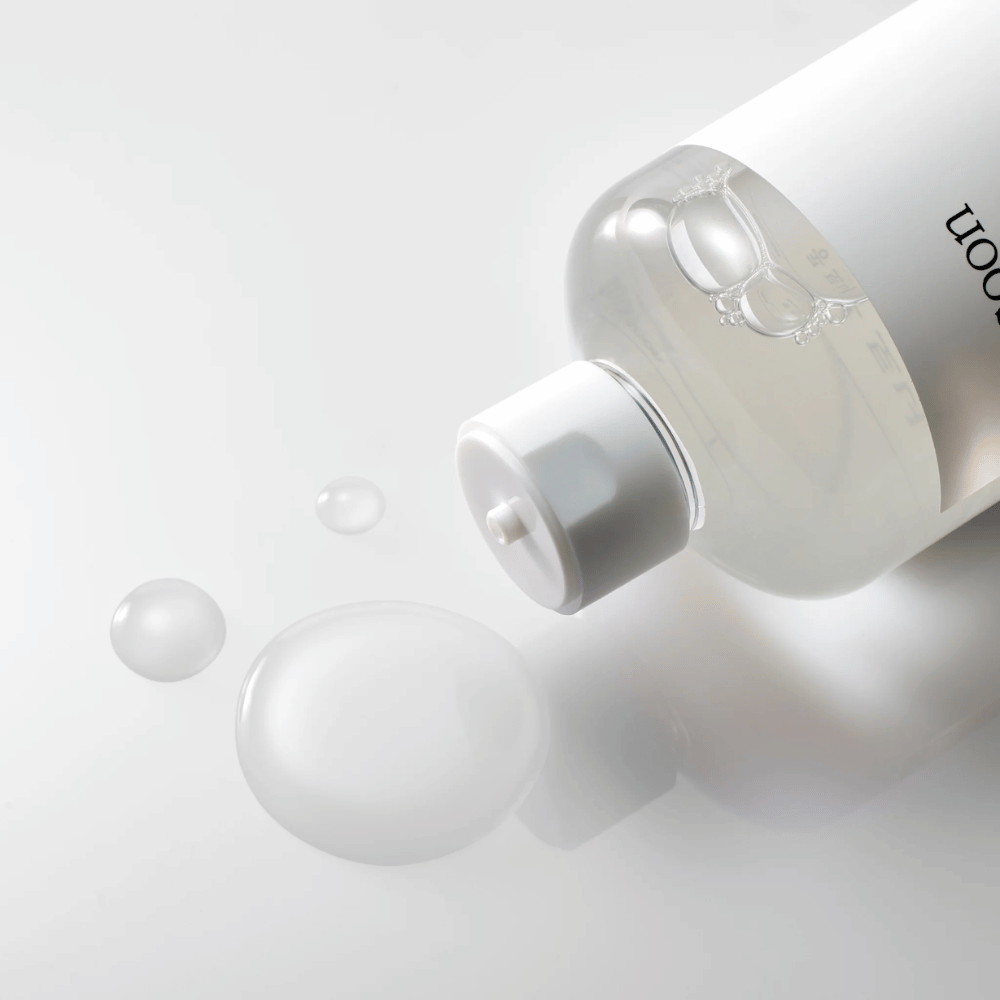 Eine durchsichtige Flasche liegt auf der Seite auf einer glänzenden Oberfläche mit einem leichten Toner, der langsam ausläuft und Tropfen sowie eine größere Pfütze auf der Oberfläche bildet. Die Flasche hat ein weißes Etikett und einen weißen Pumpmechanismus. Die Beleuchtung ist weich und schafft eine saubere und beruhigende Atmosphäre, während die leicht fließende Konsistenz und Klarheit des Toners betont wird, indem er sich auf der Oberfläche ausbreitet.