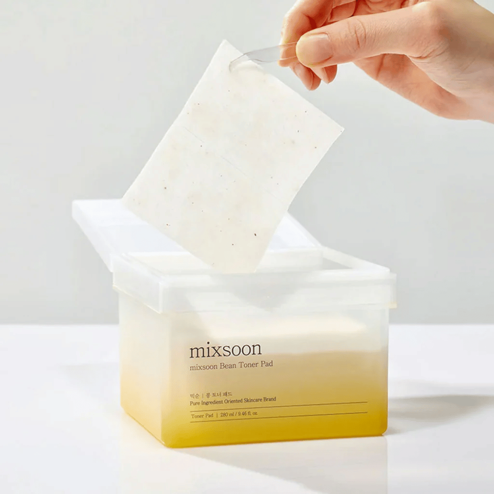 Eine Hand hält eine Pinzette und hebt ein Mixsoon Bean Toner Pad aus einem quadratischen, durchsichtigen Behälter mit einem gelblichen Ton. Der Behälter enthält 70 Toner-Pads und hat Text, der das Produkt beschreibt.
