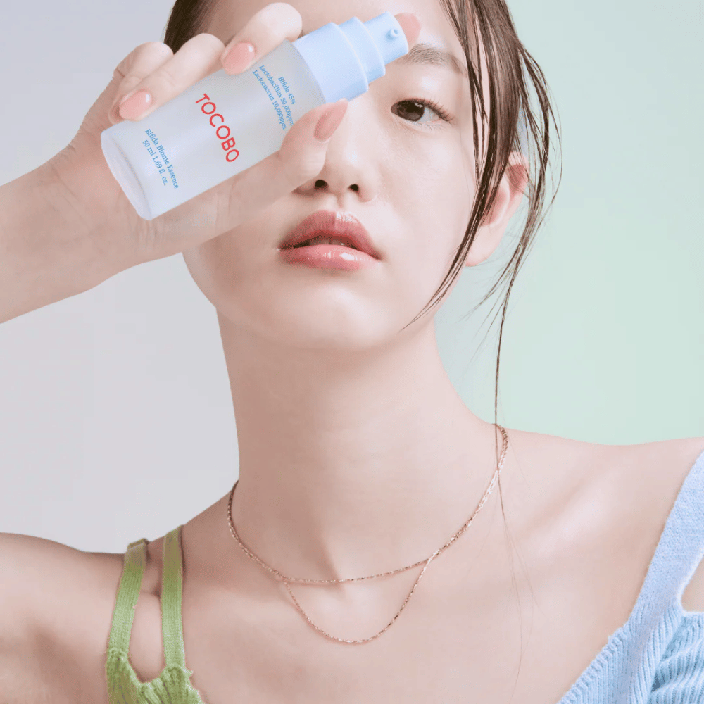 Junge Frau mit sanftem Make-up und natürlichem Hautton hält eine TOCOBO Bifida Biome Essence-Flasche vor ihrem Gesicht. Sie trägt ein hellblaues Oberteil mit grünen Trägern und eine dünne Goldkette vor einem hellen Hintergrund.