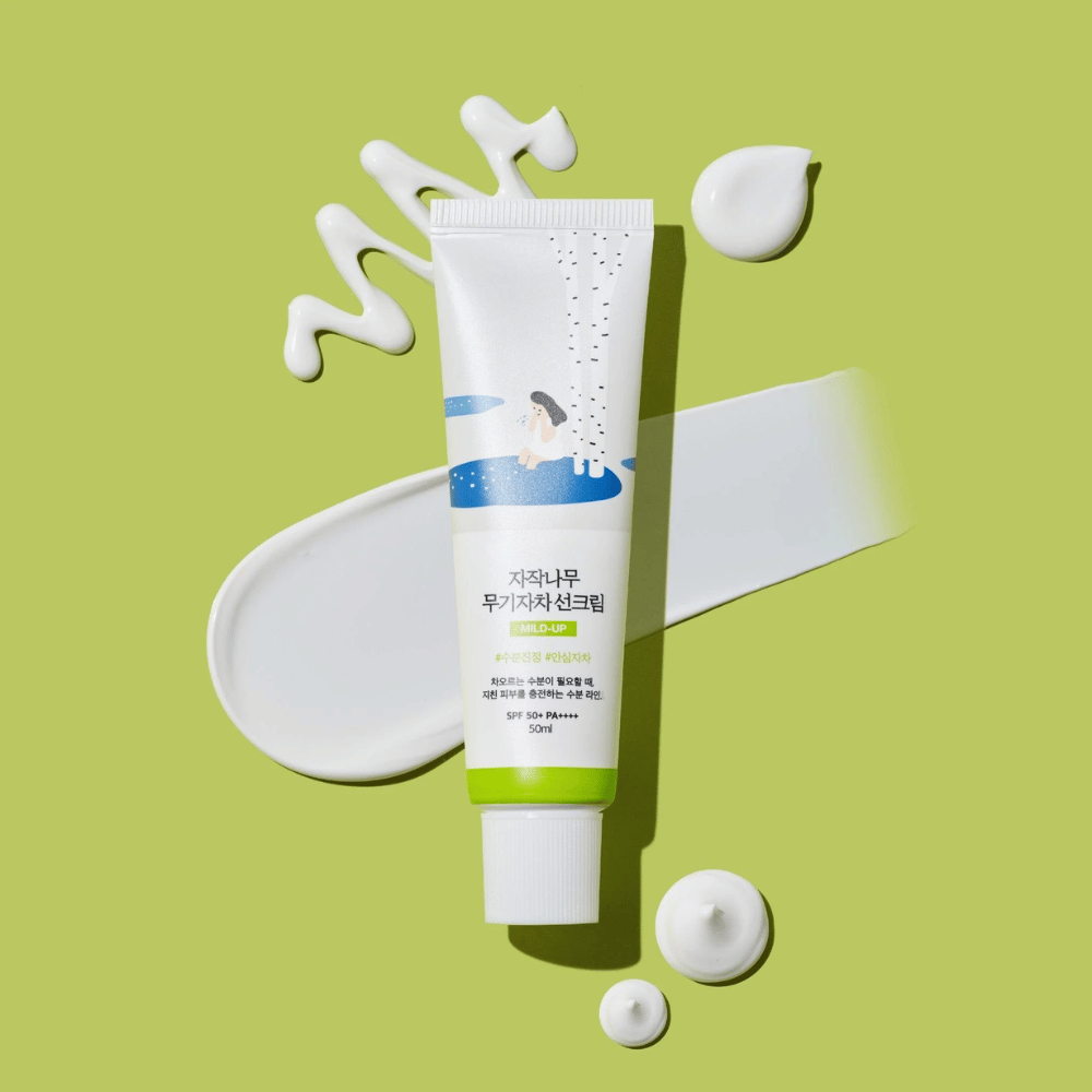 Birch Juice Moisturizing Mild Up Sunscreen – vit tub med grön bas på grön bakgrund, omgiven av utsmetad solkräm och droppar, SPF 50+ PA++++, 50 ml.