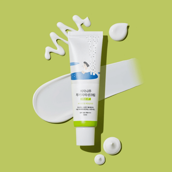 Birch Juice Moisturizing Mild Up Sunscreen – vit tub med grön bas på grön bakgrund, omgiven av utsmetad solkräm och droppar, SPF 50+ PA++++, 50 ml.