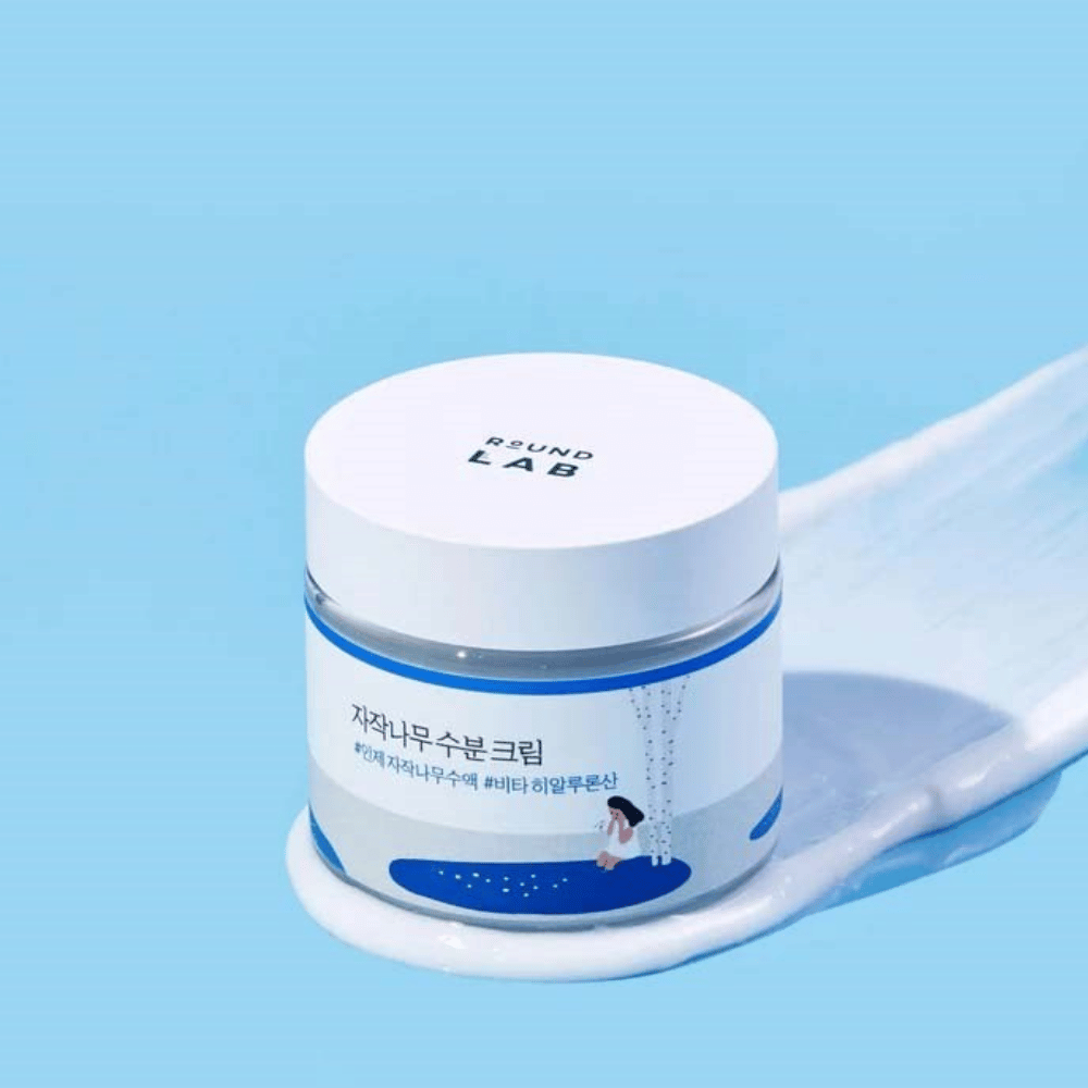 Round Lab Birch Juice Moisturizing Cream in einem bläulichen Behälter mit weißem Deckel, stehend in einer Schicht aus Creme vor blauem Hintergrund.