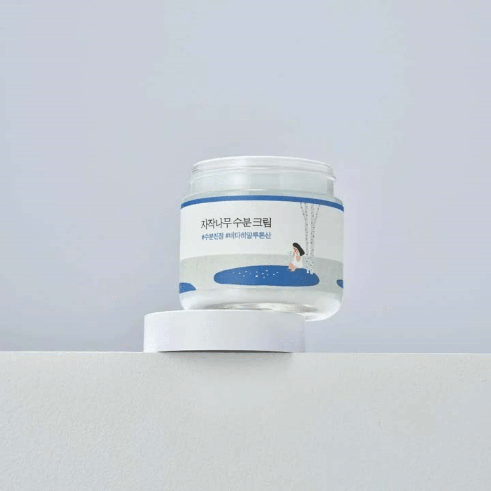 Round Lab Birch Juice Moisturizing Cream in einem transparenten Behälter mit weißem Etikett und blauer Illustration einer Frau bei einer Birke, stehend auf weißer Fläche vor hellem Hintergrund.