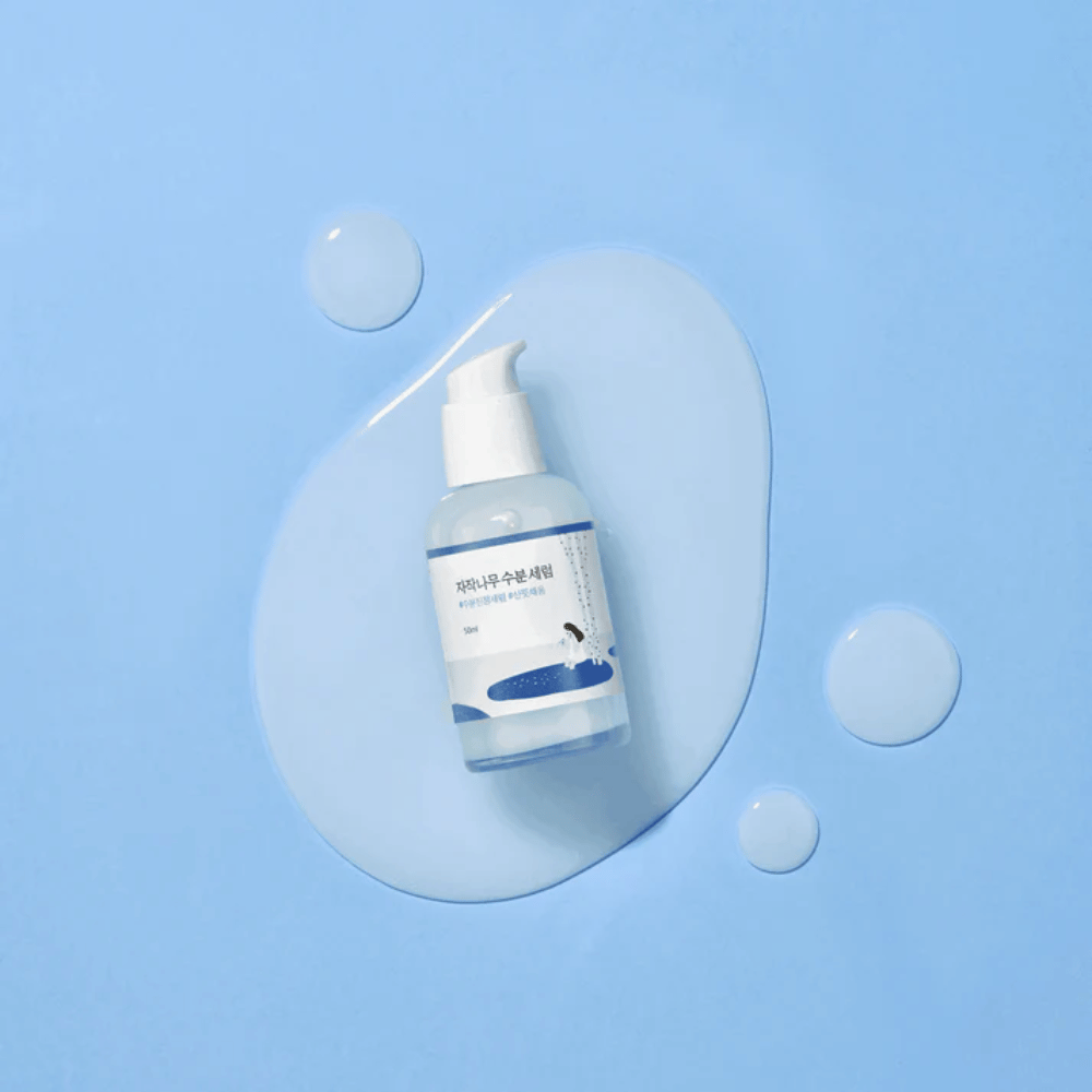 Round Lab Birch Juice Moisturizing Serum in transparenter Pumpflasche auf einem blauen Hintergrund mit Tropfen und verschüttetem Serum platziert.