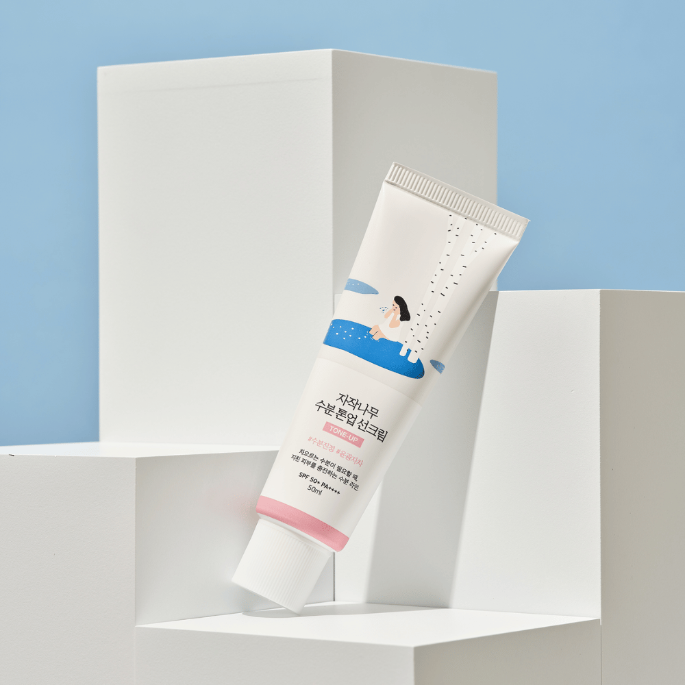 Eine Tube mit Birch Juice Moisturizing Tone Up Sunscreen platziert auf weißen Würfeln vor einem hellblauen Hintergrund. Die Tube ist weiß mit einer Illustration einer Frau bei einer Birke und Wasser. Der koreanische Text gibt an, dass das Produkt SPF 50+ PA++++ hat und 50 ml enthält.