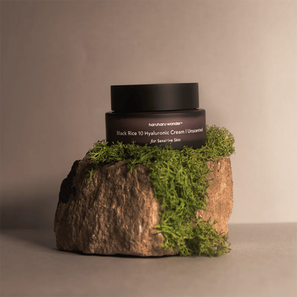 Das Bild zeigt einen Tiegel Hautcreme mit der Aufschrift "haruharu wonder Black Rice 10 Hyaluronic Cream Unscented for Sensitive Skin". Der Tiegel ist schwarz und steht auf einem großen Stein, der mit hellgrünem Moos bedeckt ist, was ein natürliches und bodenständiges Gefühl vermittelt.