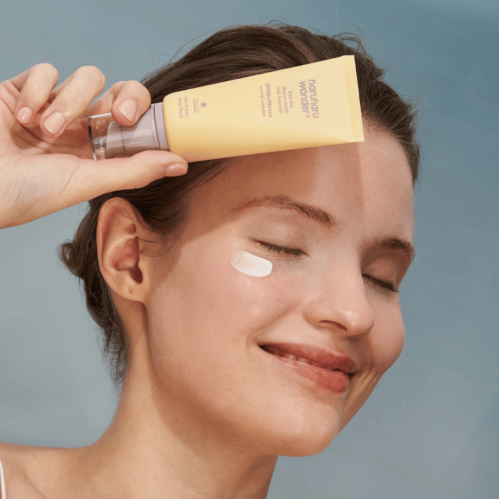 Eine lächelnde Frau trägt Black Rice Moisture Airyfit Daily Sunscreen SPF 50+ PA++++ von Haruharu Wonder auf ihr Gesicht auf. Sie hält ein gelbes Röhrchen mit Sonnencreme und hat einen Klecks des Produkts auf der Wange. Der Hintergrund ist hellblau.