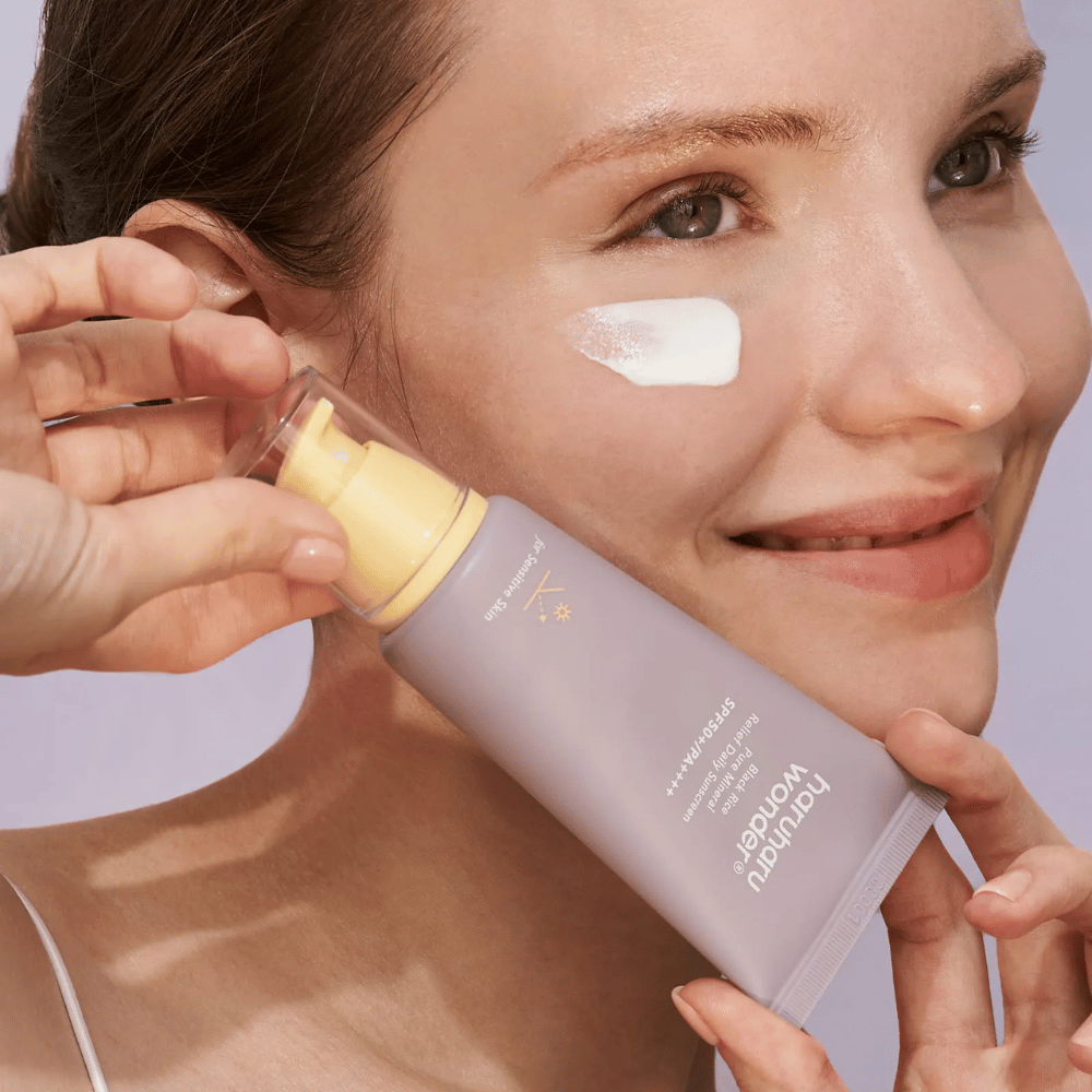 Eine lächelnde Frau trägt Haruharu Wonder Black Rice Pure Mineral Relief Daily Sunscreen SPF 50+ PA++++ auf ihr Gesicht auf. Sie hält eine lila Tube mit Sonnencreme und hat einen Klecks des Produkts auf der Wange. Der Hintergrund ist helllila.