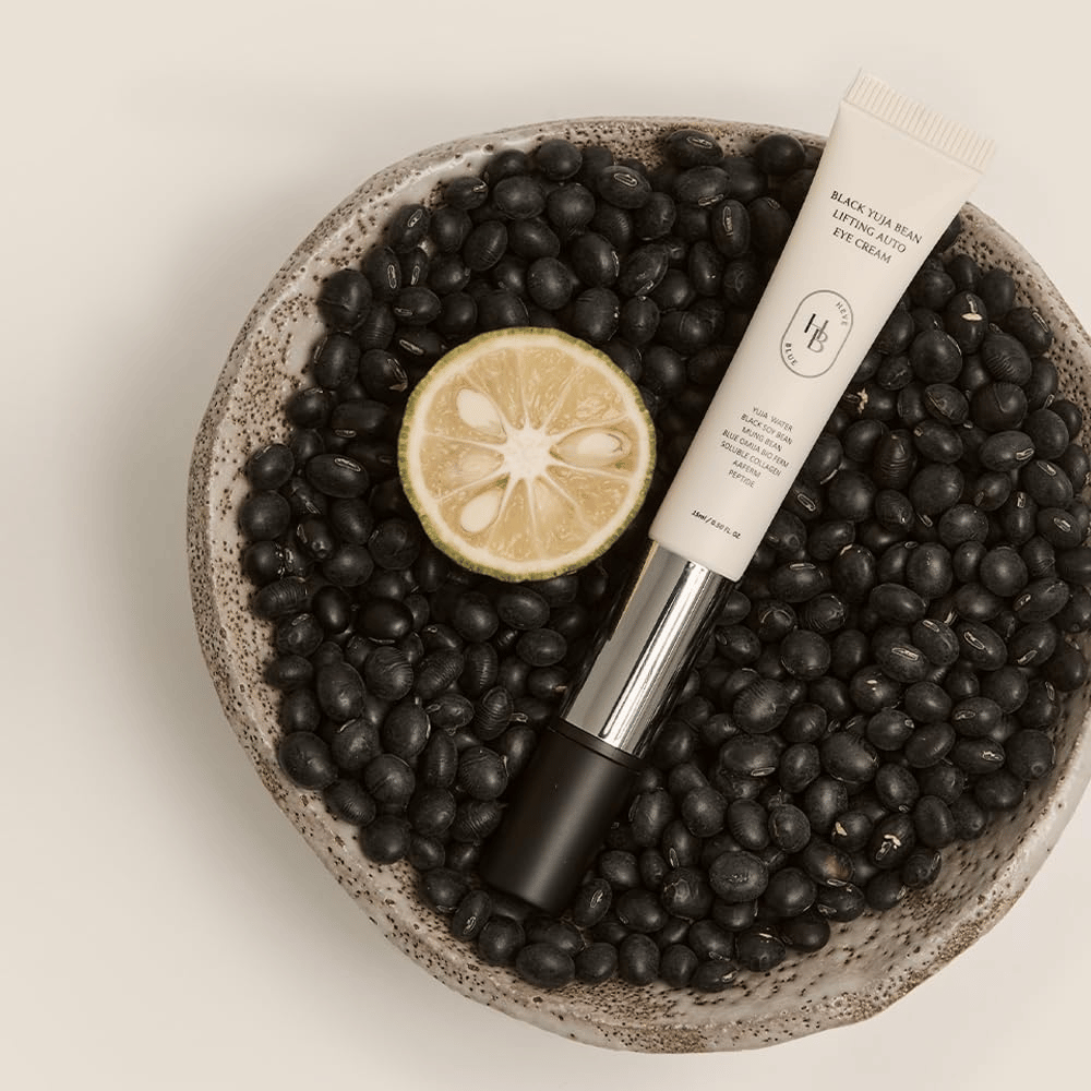 Black Yuja Bean Lifting Auto Eye Cream liegend in einer Schale gefüllt mit schwarzen Bohnen und einer halbierten Zitrusfrucht – eine visuelle Darstellung der natürlichen Inhaltsstoffe des Produkts.