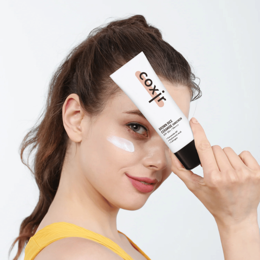 Frau mit auf die Wange aufgetragener Sonnencreme hält eine Tube Coxir Brown Rice Ceramide Sunscreen, SPF 50+ PA+++, zur Stirn und lächelt leicht.