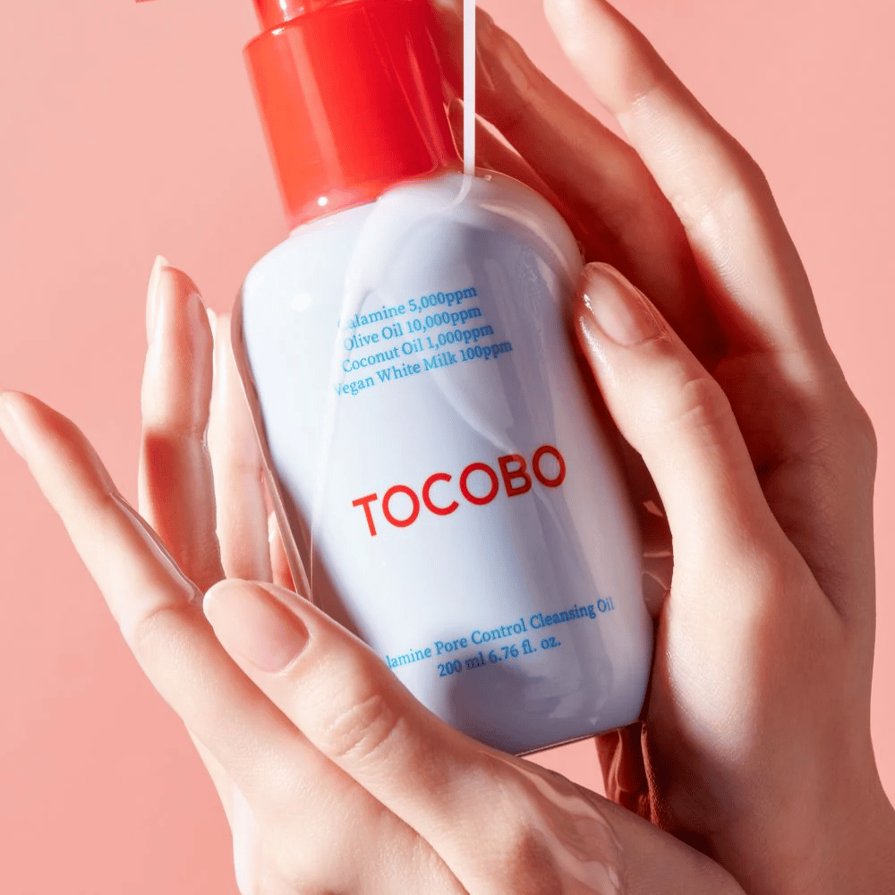 TOCOBO Calamine Pore Control Cleansing Oil wird in Händen vor einem rosa Hintergrund gehalten, die rote Pumpe der Flasche und die Zutatenliste sind deutlich sichtbar.