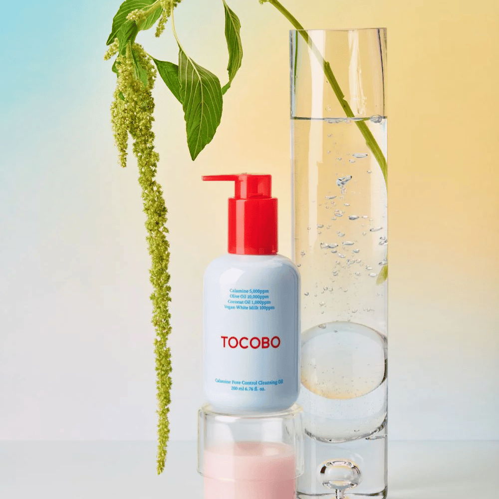 TOCOBO Calamine Pore Control Cleansing Oil mit roter Pumpe steht neben einem hohen Glasrohr, gefüllt mit Wasser und einer grünen Pflanze, vor einem hellen pastellfarbenen Hintergrund.