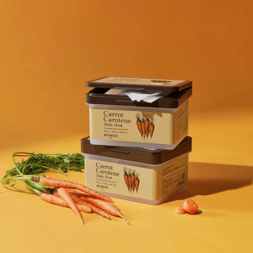 Das Bild zeigt drei gestapelte Verpackungen von "Carrot Carotene Daily Mask" von SKINFOOD, platziert auf einem orangefarbenen Hintergrund zusammen mit frischen Karotten. Jede Schachtel enthält Gesichtsmasken, die mit β-Carotin aus Karottensamenöl angereichert sind, für den täglichen Gebrauch konzipiert, um die Haut zu nähren und zu pflegen.