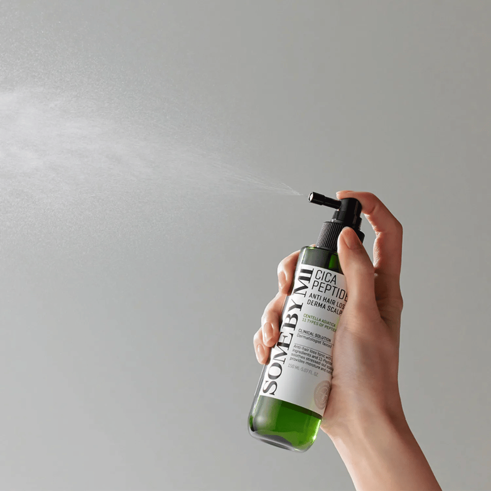 Hand, die SOME BY MI Cica Peptide Anti Hair Loss Derma Scalp Tonic aus einer grünen Flasche mit schwarzem Sprühkopf und weißem Etikett gegen grauen Hintergrund sprüht.