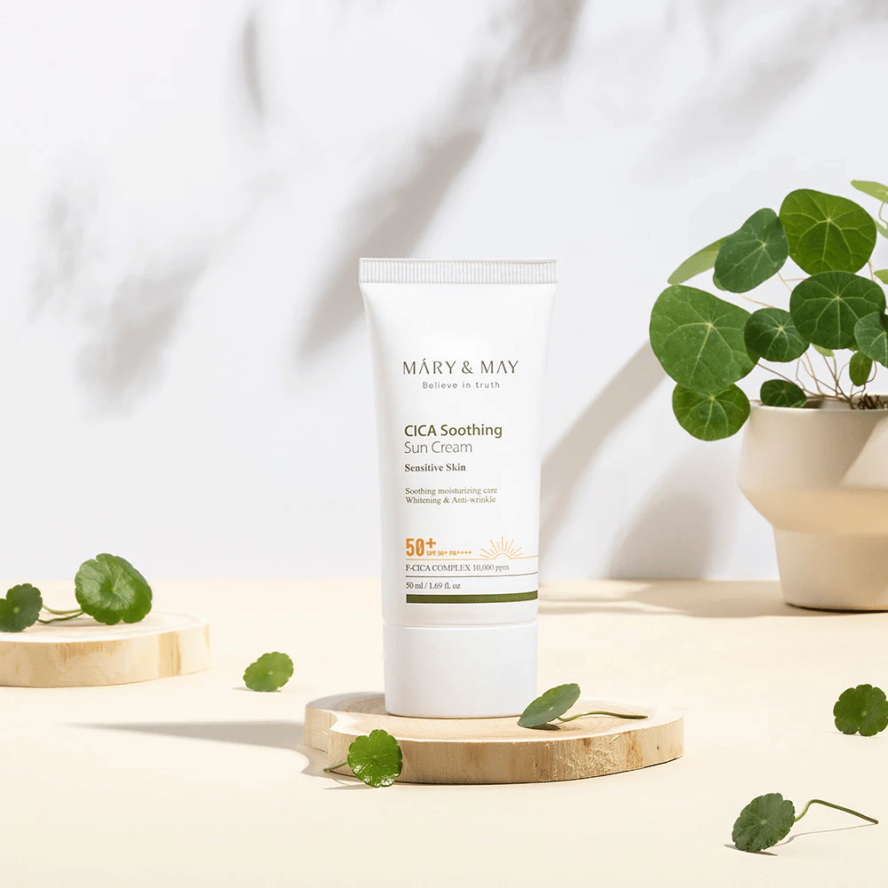 Mary&May CICA Soothing Sun Cream auf einem Holzpodest platziert, umgeben von grünen Centella-Blättern. Das Bild betont die hautberuhigenden Inhaltsstoffe und die natürliche Inspiration hinter dem Produkt.