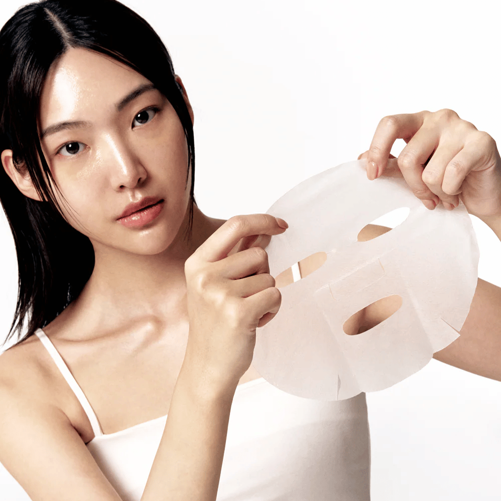 Frau mit sauberer und mit Feuchtigkeit versorgter Haut hält eine weiße Sheet mask vor sich, bereit, sie auf das Gesicht aufzutragen.