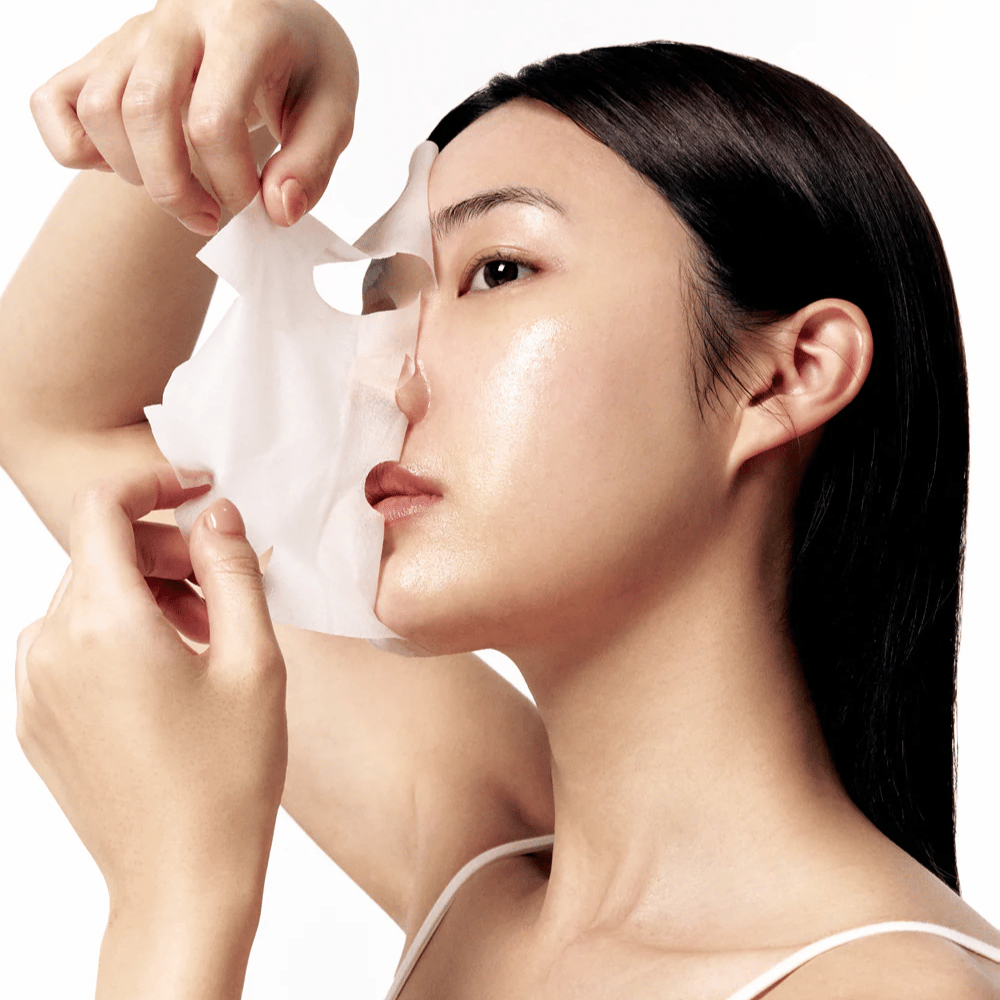 Frau mit glatter, mit Feuchtigkeit versorgter Haut nimmt eine weiße Sheet mask vom Gesicht ab, was einen strahlenden und gleichmäßigen Hautton enthüllt.