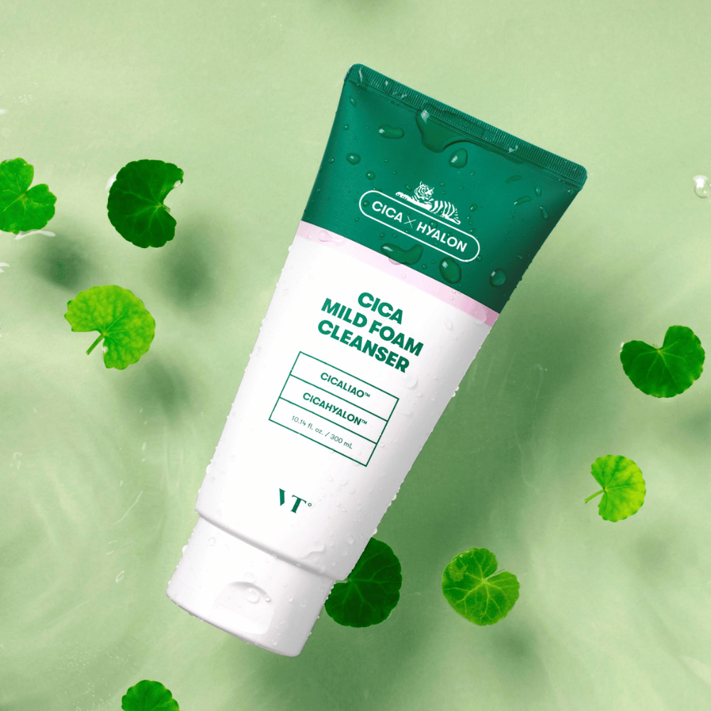 VT Cica Mild Foam Cleanser liegend auf einem hellgrünen Untergrund mit Wassertropfen auf der Verpackung und umgeben von grünen Centella Asiatica-Blättern, was die beruhigenden Eigenschaften des Produkts hervorhebt.