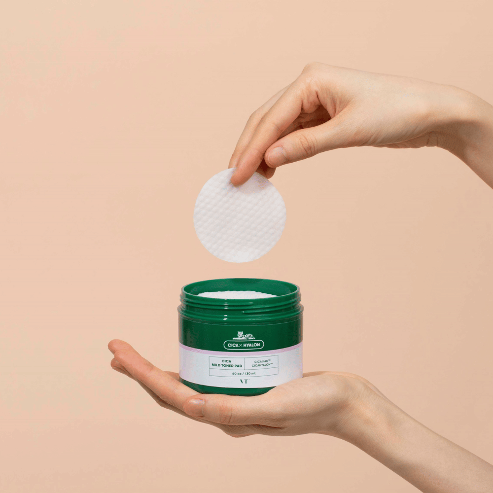 Hand, die eine grüne Dose mit VT Cica Mild Toner Pad hält und ein rundes, weißes Tonerpad über der Dose vor beigem Hintergrund anhebt.