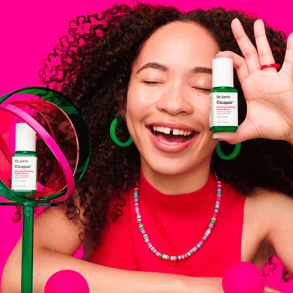 Eine Frau lächelt breit, während sie Dr.Jart+ Cicapair Intensive Soothing Repair Serum vor ihr Gesicht hält. Sie trägt grüne Ohrringe und eine rote Halskette. Der Hintergrund ist hellrosa und mit grünen und rosa Kreisen dekoriert.