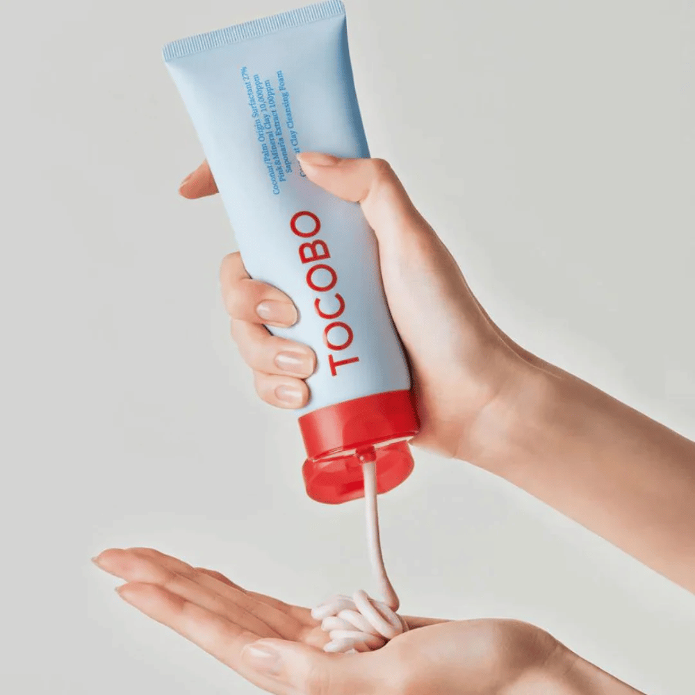 Eine Person hält eine Tube von TOCOBO Coconut Clay Cleansing Foam. Die Tube ist hellblau mit einem roten Deckel und dem Text "TOCOBO" auf der Vorderseite. Das Produkt wird aus der Tube gedrückt und formt sich wie ein Shampoo in der Hand. Der Hintergrund ist neutral und lenkt den Fokus auf das Produkt.
