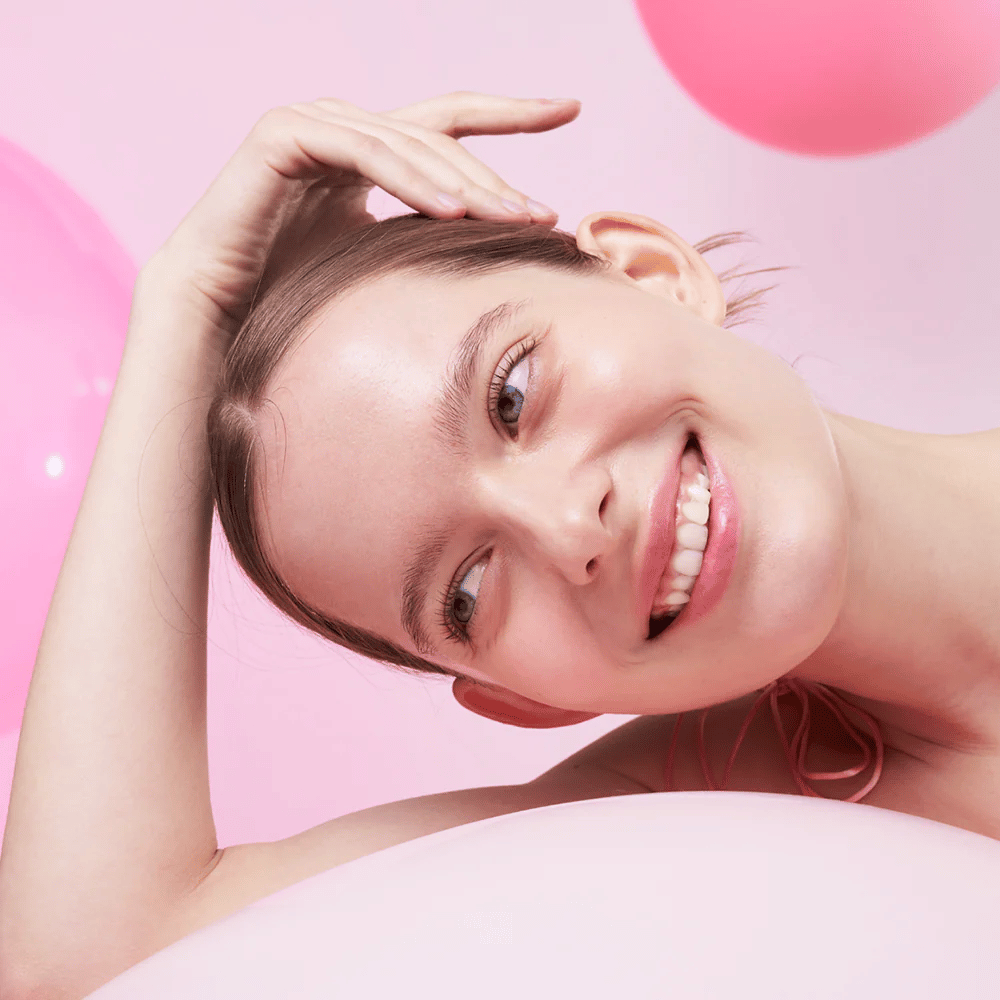 Eine Frau lächelt und ruht ihren Kopf auf der Hand, umgeben von rosa Luftballons. Ihre Haut sieht strahlend und natürlich gesund aus.