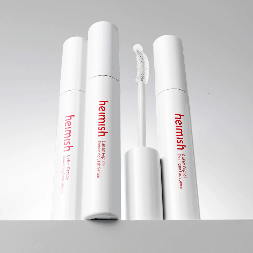 Mehrere Verpackungen von Heimish Dailism Peptide Enhancing Lash Serum werden zusammen mit der gebogenen Applikatorbürste gezeigt. Das Bild hebt das elegante Design des Produkts und die Präzisionsbürste für ein einfaches Auftragen hervor.