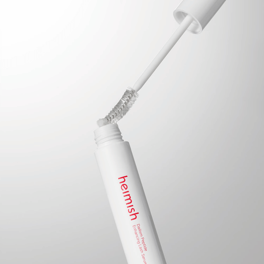 Nahaufnahme des Applikators von Heimish Dailism Peptide Enhancing Lash Serum. Die Bürste wird aus dem weißen Behälter herausgezogen und zeigt die transparente Serumformel, die auf die Wimpern aufgetragen wird.