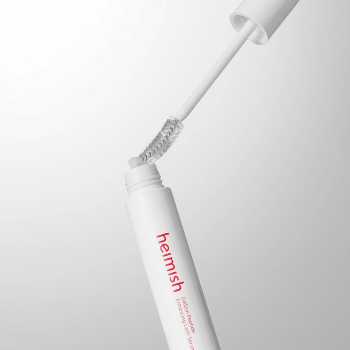 Nahaufnahme des Applikators von Heimish Dailism Peptide Enhancing Lash Serum. Die Bürste wird aus dem weißen Behälter herausgezogen und zeigt die transparente Serumformel, die auf die Wimpern aufgetragen wird.