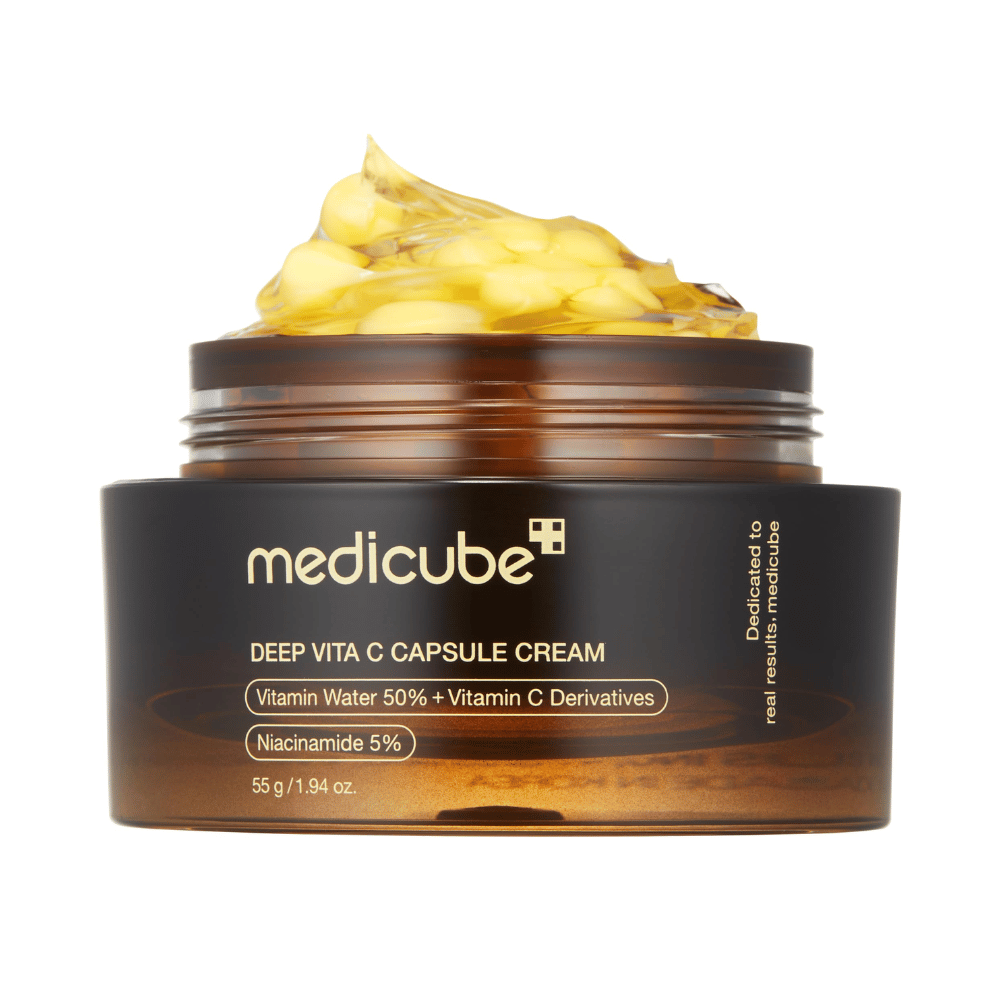 Eine Nahaufnahme von "Medicube Deep Vita C Capsule Cream", die die Konsistenz der Creme und die Vitamin-C-Kapseln zeigt. Die Verpackung ist dunkelbraun mit goldener Schrift und beschreibt den Inhalt von Vitaminwasser (50 %) und Niacinamid (5 %), was eine visuelle Darstellung der feuchtigkeitsspendenden und hautverbessernden Eigenschaften des Produkts bietet.