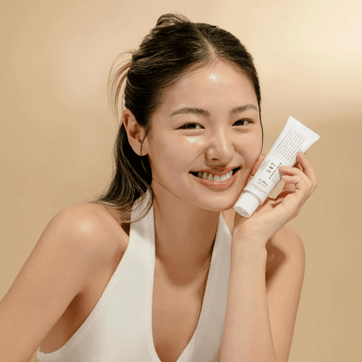 Modell med glowig hud som ler och håller i Dr. Althea 147 Barrier Cream – en koreansk ansiktskräm för hudbarriär och återfuktning.