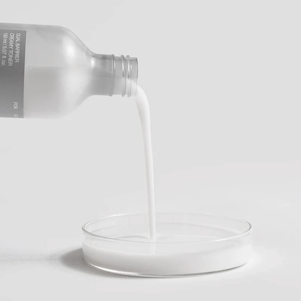 Celimax Dual Barrier Creamy Toner wird vorsichtig aus der Flasche in einen runden Laborbrunnen gegossen, wobei die milchige und nährstoffreiche Formel deutlich im Fokus steht.