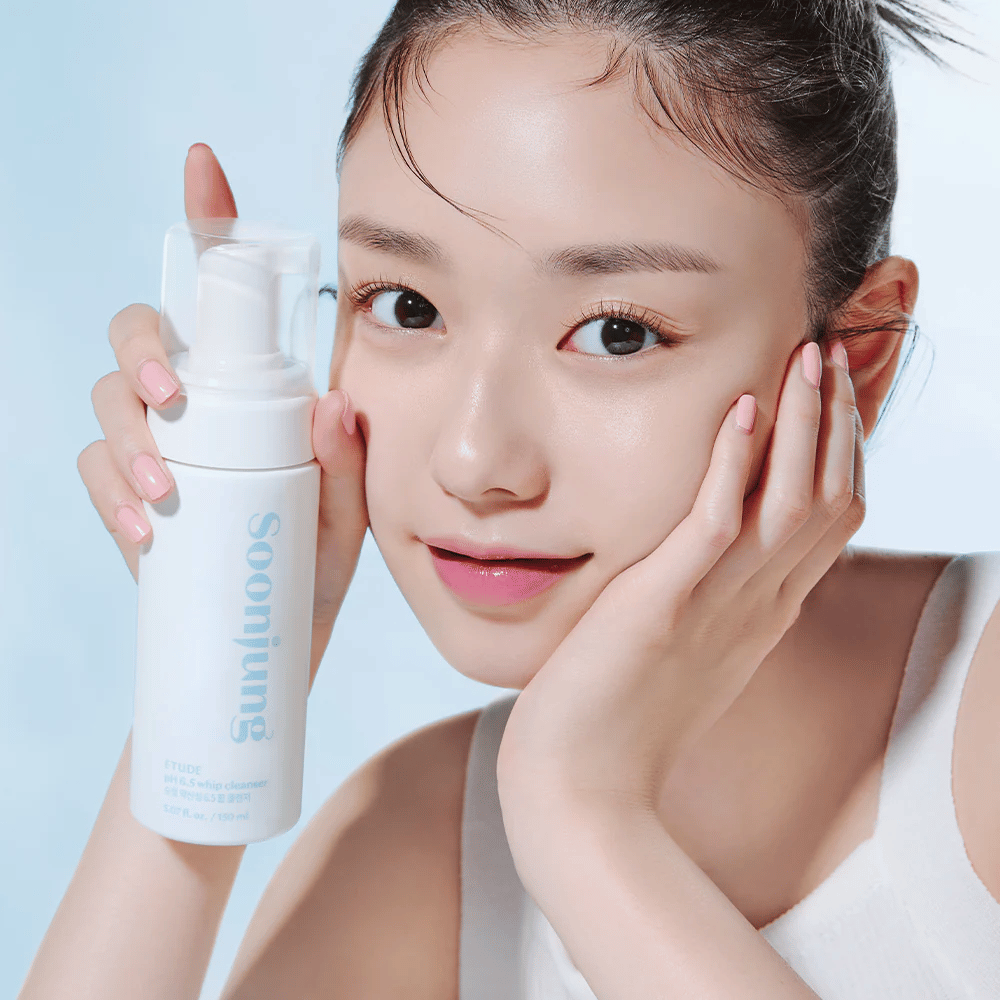 Eine junge Frau mit klarer Haut hält eine weiße Pumpflasche mit dem Text "Soon Jung pH 6.5 Whip Cleanser" von ETUDE. Sie lächelt in die Kamera mit leichtem Make-up und einer frischen Ausstrahlung vor einem hellblauen Hintergrund.