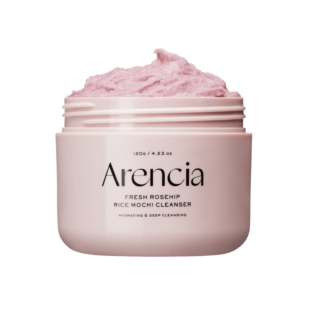 Nahaufnahme eines rosa Behälters mit Arencia Royal Rosehip Cleanser, gefüllt mit rosa Reinigungscreme mit einer geschlagenen, mochi-ähnlichen Textur. Der Text auf dem Behälter zeigt, dass das Produkt feuchtigkeitsspendend ist und tiefenreinigt.