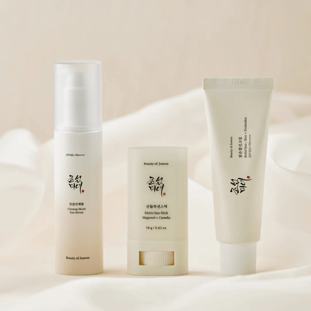 Drei Sonnenschutzprodukte von Beauty of Joseon – Ginseng Moist Sun Serum, Matte Sun Stick und Relief Sun Rice + Probiotics – stehen aufgereiht auf einem cremefarbenen Stoff mit weichem Licht.