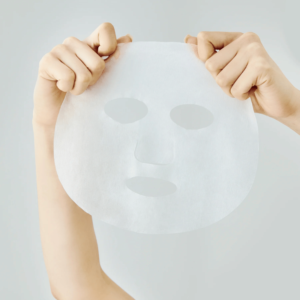 Zwei Hände halten eine weiße Blattmaske vor einem hellgrauen Hintergrund hoch, mit sichtbaren Ausschnitten für Augen, Nase und Mund.