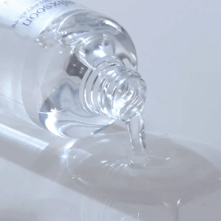 Närbild på genomskinligt serum som hälls ur en flaska Mixsoon Glacier Water Hyaluronic Acid Serum på en blank yta.