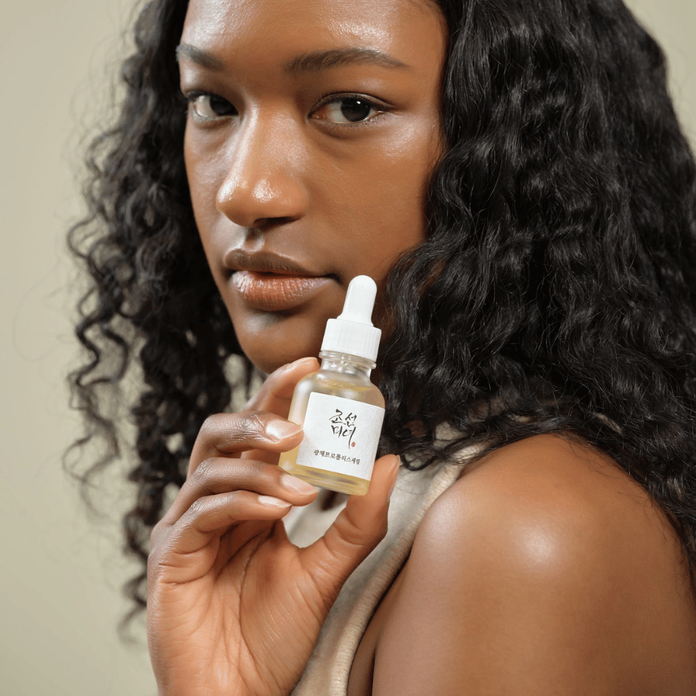 Frau mit lockigem Haar hält eine Flasche Beauty of Joseon Glow Serum mit Pipette, ein feuchtigkeitsspendendes und strahlendes Hautpflegeprodukt.