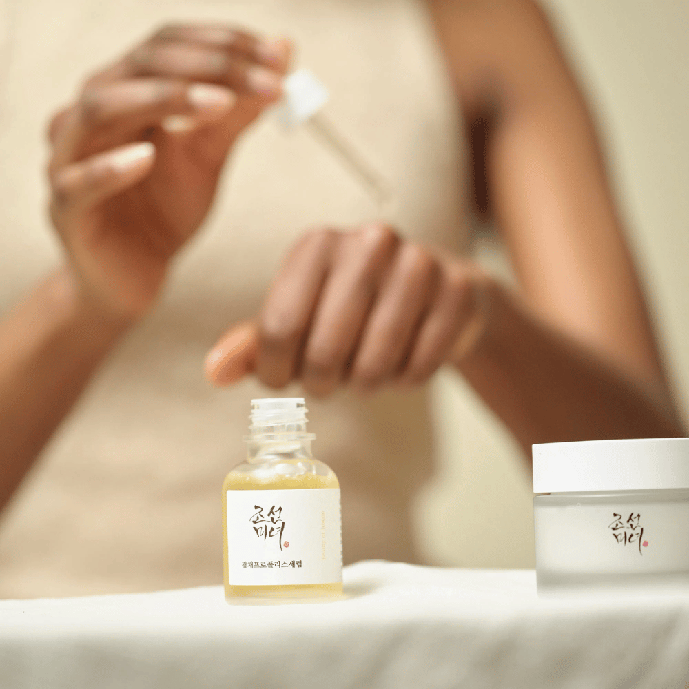 Person, die Beauty of Joseon Glow Serum mit Pipette aus Glasflasche aufträgt, mit einem Glas derselben Serie daneben auf dem Tisch.